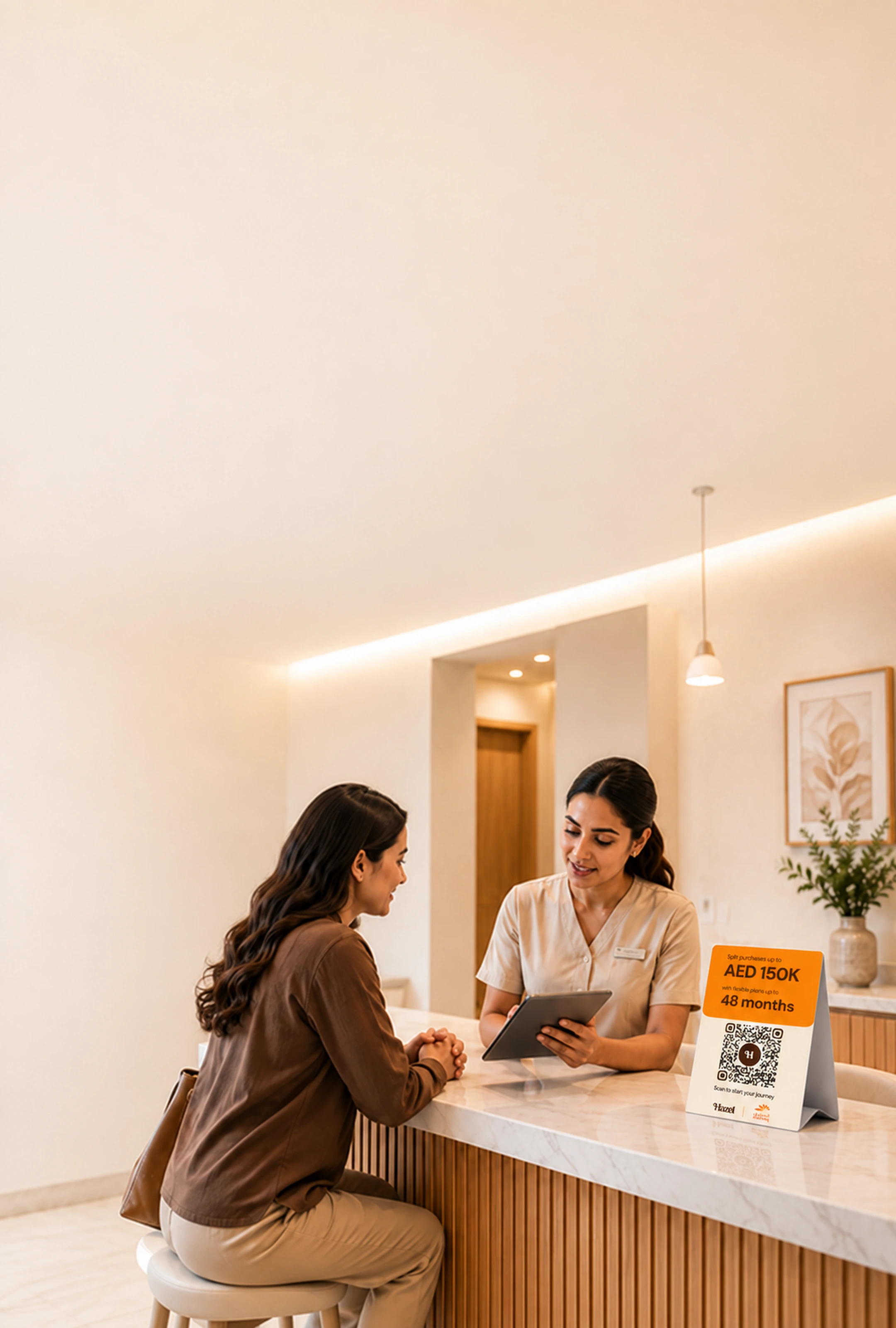 A clinic team member reviews a tablet with a seated patient at the reception counter, a Hazel payment card showing AED 150K over 48 months beside them.