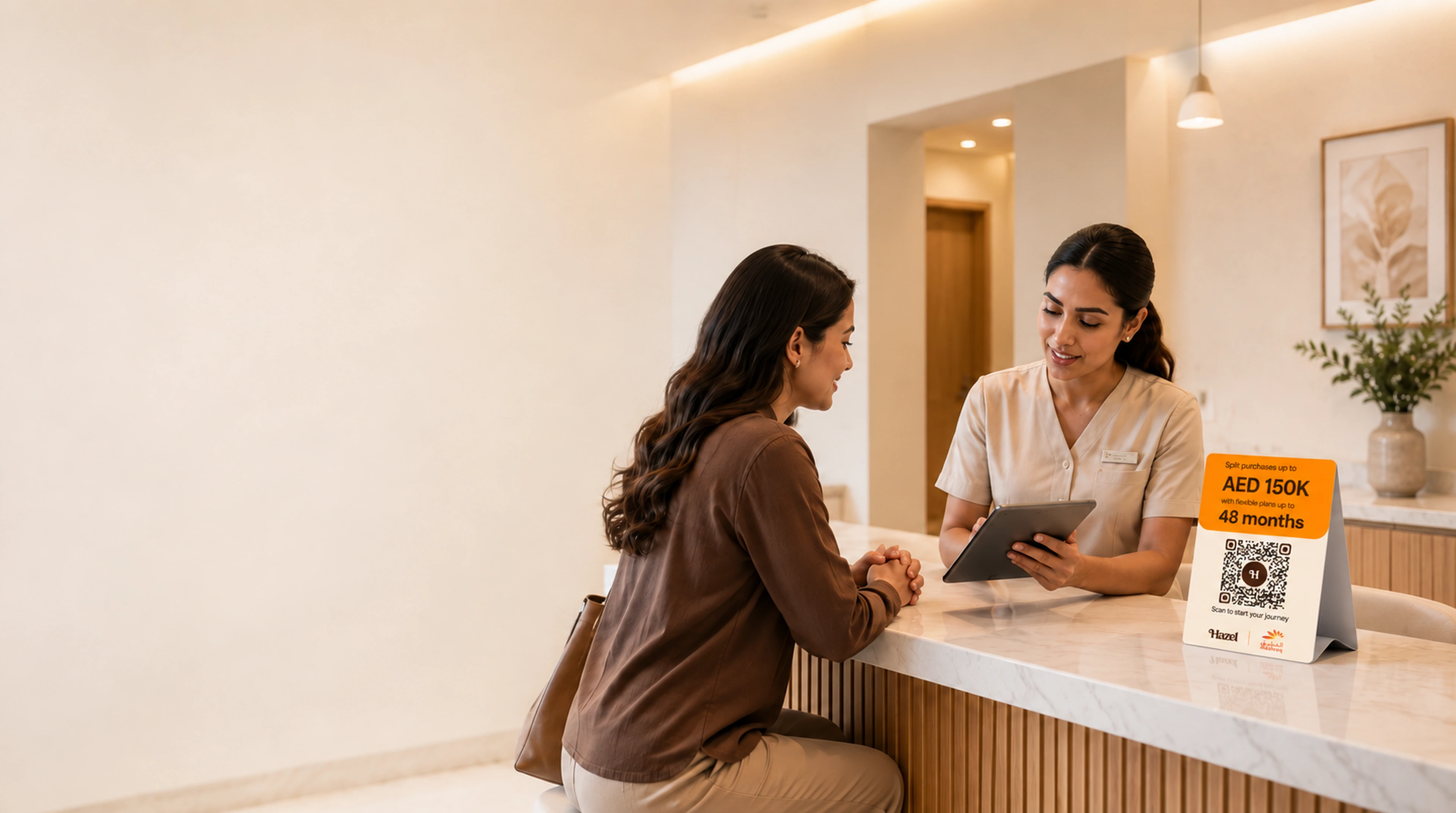 A clinic team member reviews a tablet with a seated patient at the reception counter, a Hazel payment card showing AED 150K over 48 months beside them.