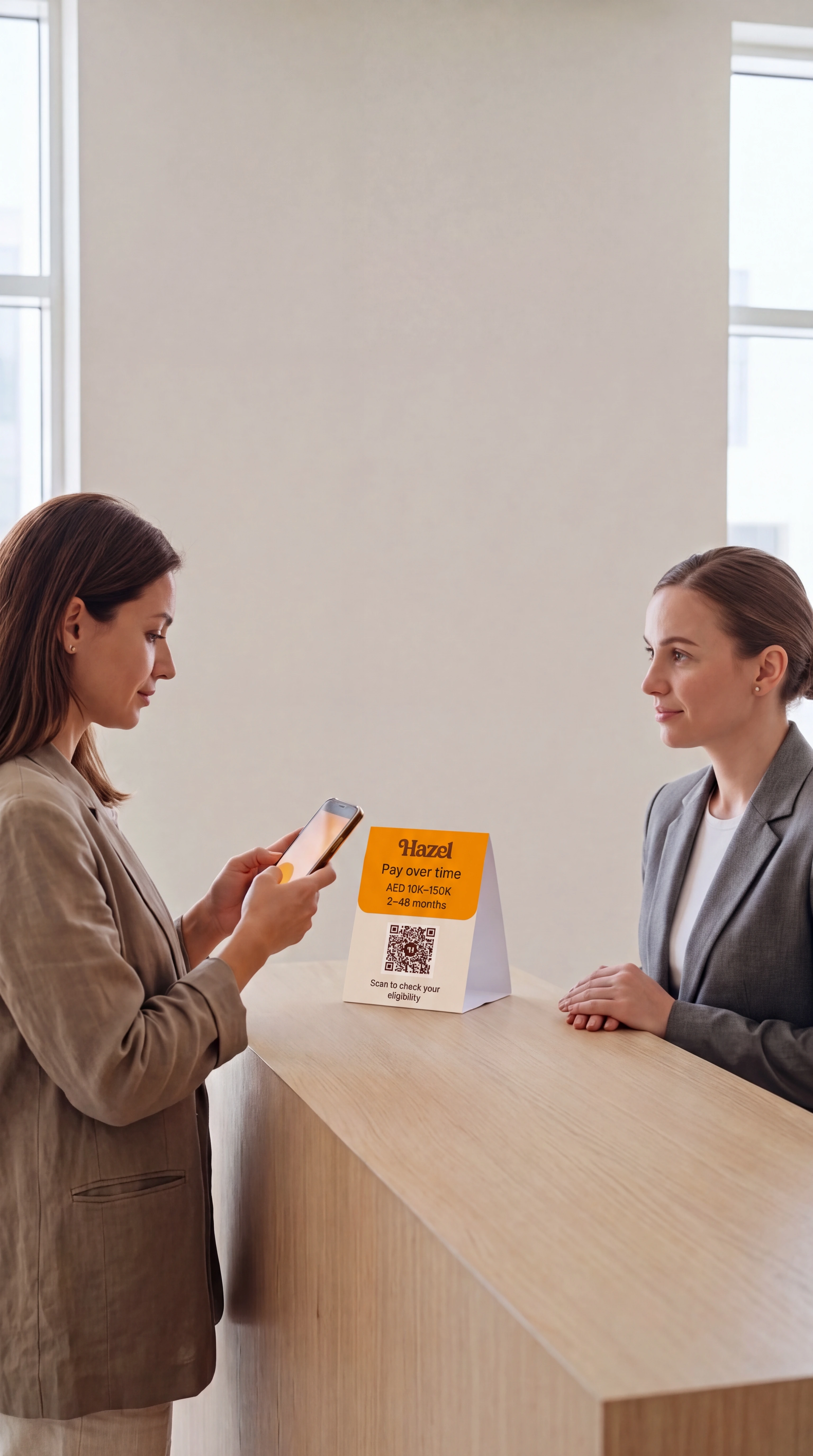 A patient at a sunlit clinic reception scans a Hazel QR card with her phone while the receptionist opposite her looks on.