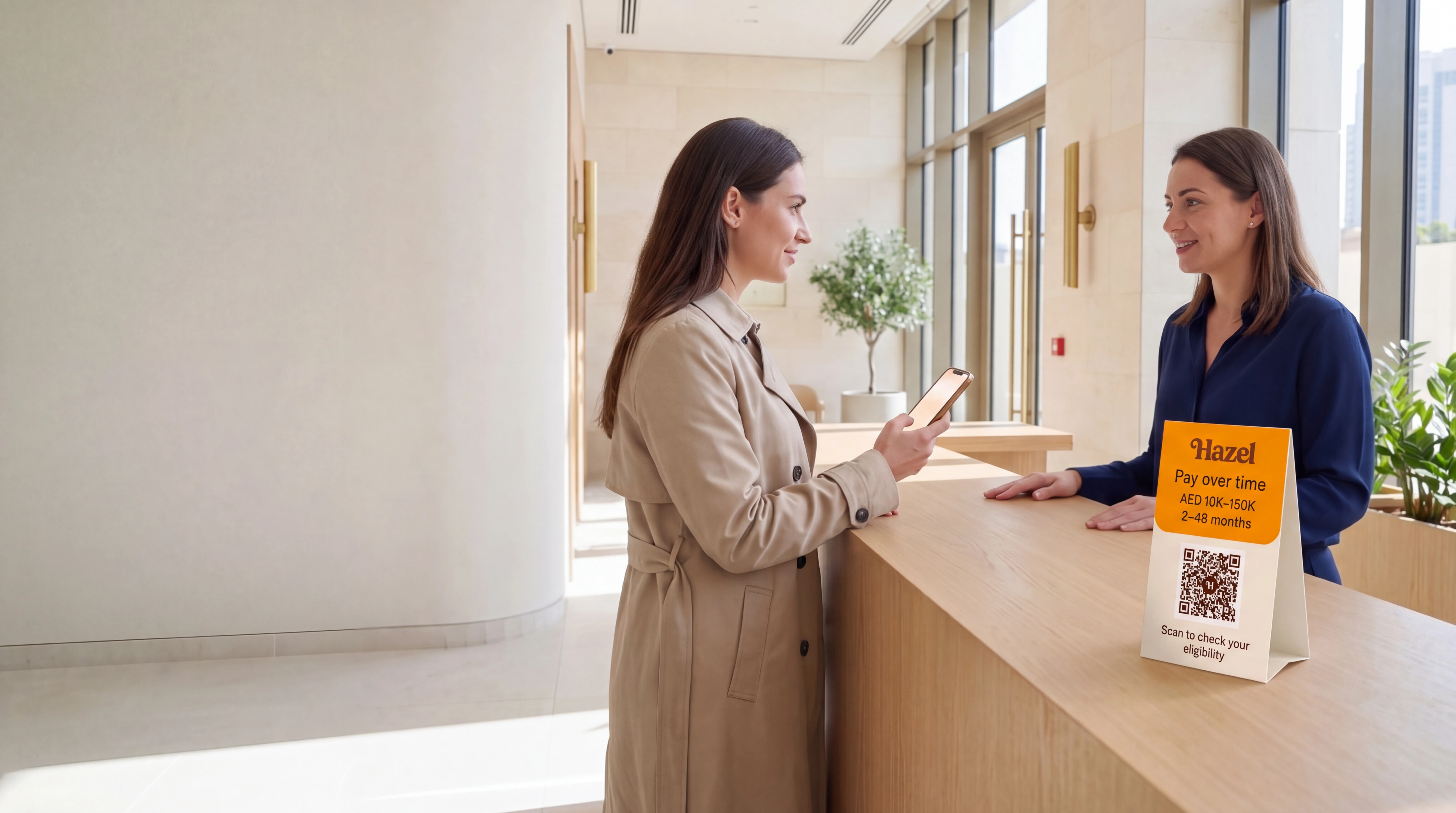 A patient at a sunlit clinic reception scans a Hazel QR card with her phone while the receptionist opposite her looks on.